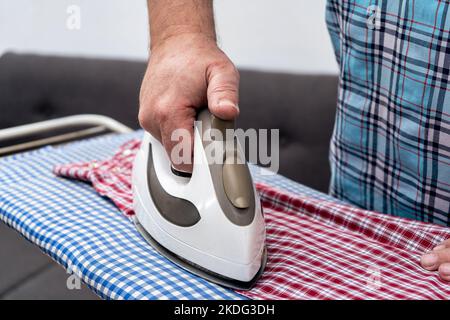 Nahaufnahme der Hand eines Mannes, der ein Bügeleisen hält, während er ein Hemd auf einem Brett bügelt. Konzept der Hausarbeit, Hausarbeit. Horizontaler Rahmen. Stockfoto