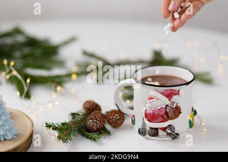 Weibliche Hand gießt Marschwalben in eine weiße Tasse heißen Kakaos auf einem weißen Tisch mit einem Weihnachtsbaum. Stockfoto