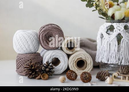 Eine Komposition mit Kugeln aus Fäden verschiedener Farben, Kegeln, Schlitten, einem Glas, das mit Macrame auf einem weißen Tisch verziert ist Stockfoto