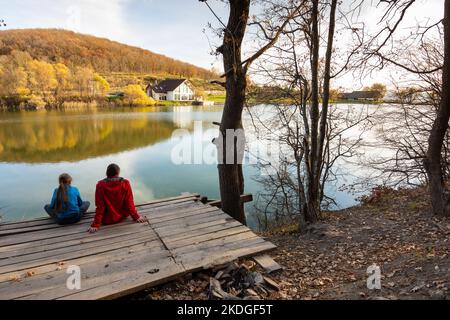 Ein Mädchen und ein Mädchen sitzen auf einer Holzbrücke am See und blicken auf eine wunderschöne Herbstlandschaft Stockfoto