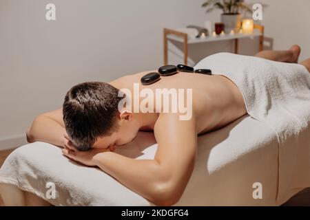 Schöner Mann, der sich während einer Rückenmassage mit heißen Steinen entspannt. Körperpflege und SPA im Resort. Stockfoto