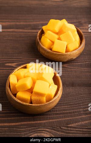 Getrocknete und kandierte Mangowürfel in Holzschüsseln auf braunem, strukturiertem Holzhintergrund. Seitenansicht, Nahaufnahme, vegan, vegetarisches Food-Konzept. Stockfoto