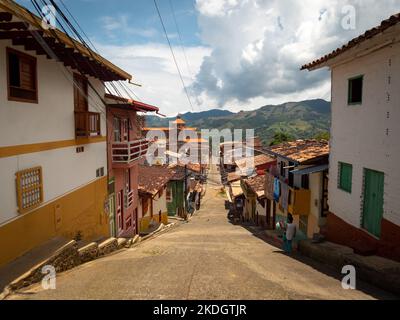 Jerico, Antioquia, Kolumbien - April 5 2022: Schmale Kolonialstraße mit bunten Häusern und Kathedrale aus orangefarbenem Backstein vor dem Hintergrund üppiger Berge Stockfoto