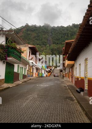 Jerico, Antioquia, Kolumbien - April 5 2022: Graue Brick Street mit bunten Häusern vor üppigem Berggrund mit Wasserfall Stockfoto