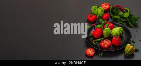 Grüne und rote gerippte Tomaten, amerikanische oder florentinische Sorte. Reifes Gemüse, Chili, frische Lorbeerblätter. Trendiges hartes Licht, dunkler Schatten, Steinbeton Stockfoto