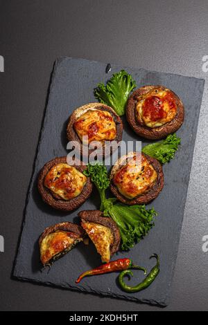 Gebackene gefüllte Pilzkappen bereit zum Essen. Champignons, Gewürze, Gemüse, Käse. Servierte Portion, trendiges hartes Licht, dunkler Schatten. Schwarzer Stein-Kon Stockfoto