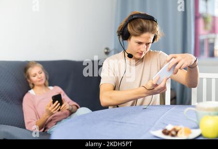 Junge, der am Telefon ein Spiel spielt, Mutter wartet auf ihn Stockfoto