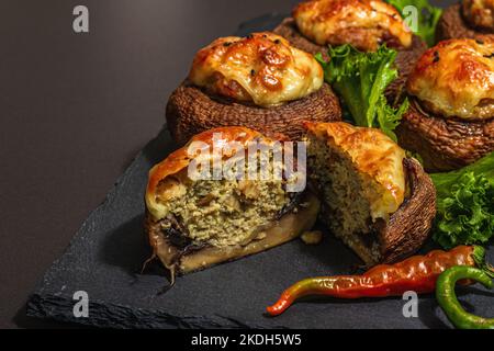 Gebackene gefüllte Pilzkappen bereit zum Essen. Champignons, Gewürze, Gemüse, Käse. Servierte Portion, trendiges hartes Licht, dunkler Schatten. Schwarzer Stein-Kon Stockfoto