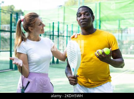 Mann und Frau mit Schlägern in den Händen plaudern, nachdem sie Padel auf dem Tennisplatz gespielt haben Stockfoto