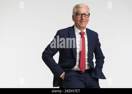 Ein Geschäftsmann mit einem Lächeln auf dem Gesicht, der seine Hände in die Taschen legte und vor grauem Hintergrund saß Stockfoto