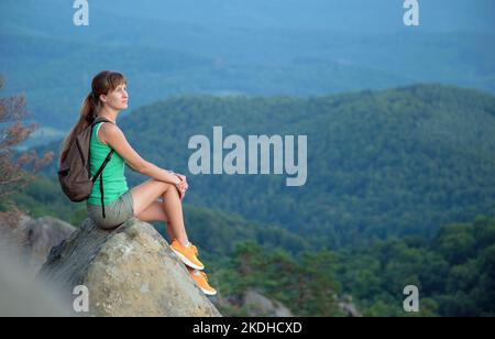 Sportliche Wanderin, die alleine auf einem Bergwanderweg eine Pause einnimmt. Einsame Frau, die den Blick auf die abendliche Natur von den felsigen Klippen auf dem Wildnispfad genießt Stockfoto