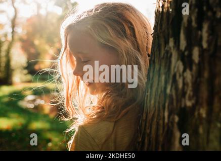 Portrait eines Mädchens, das bei Sonnenuntergang versteckt und hinter einem Baum sucht Stockfoto