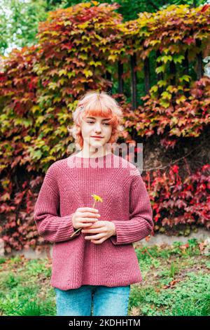 Junge rothaarige Mädchen, die gegen bunte Herbstlaub stehen und gelbe Blume halten. Stockfoto