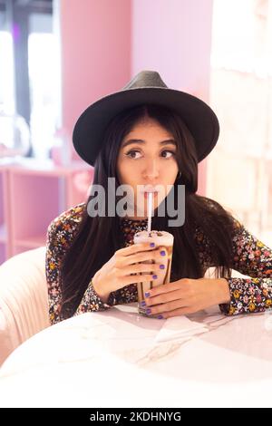 Genießen Sie einen Drink mit Sorbet eine junge lateinerin mit Hut und langen Haaren, entspannter Lebensstil im Restaurant, Modell in gemustertem Kleid Stockfoto