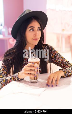 Sitzende junge Frau mit Hut und gemustertem Kleid mit Glas kalter Schokolade Getränk mit Sorbet, latin Modell Lifestyle, Schönheit Stockfoto