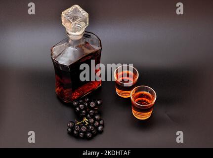 Hausgemachter Apfelbeerlikör auf schwarzem Hintergrund, ein paar reife Beeren, eine Glasflasche und zwei Gläser. Nahaufnahme. Stockfoto