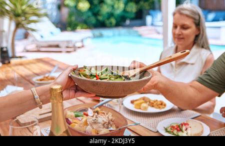 Hände, Essen und Familie entspannen sich auf einer Terrasse, essen, binden und teilen sich eine Mahlzeit an einem Tisch zusammen. Freunde, Salat und ältere Frau ruhig, zufrieden und genießen Stockfoto