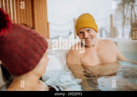 Das ältere Paar mit kitschiger Kappe genießt an kalten Wintertagen gemeinsam die Badewanne im Freien auf der Terrasse. Stockfoto