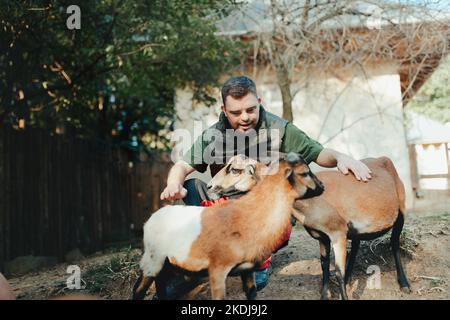 Hausmeister mit Down-Syndrom kümmert sich um Tiere im Zoo, streichelt Ziegen. Konzept der Integration von Menschen mit Behinderungen in die Gesellschaft. Stockfoto