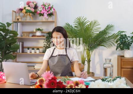 Floristenkonzept, weibliche Floristin prüft Blumenbestellungen auf Laptop und notiert sich im Notebook im Geschäft. Stockfoto