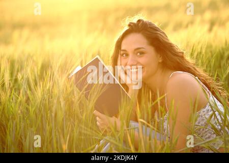 Ein glücklicher Leser, der ein Buch hält, schaut bei Sonnenuntergang in einem Weizenfeld auf die Kamera Stockfoto