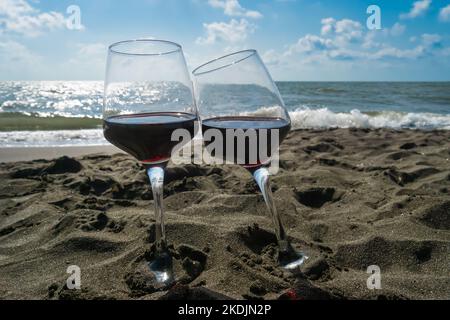 Nahaufnahme von zwei Gläsern Rotwein, die im Sand am Strand vor einem blauen Himmel mit Wolken und Sonnenstrahlen stehen Stockfoto