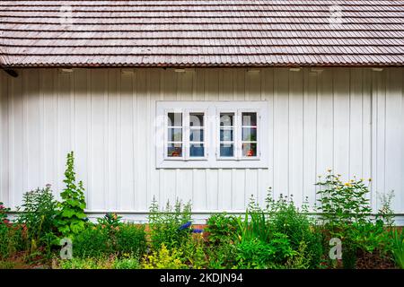 Vorderansicht der Fassade des alten Hauses. Weiß lackierte Bretter und ein Schinddach. Wunderschöner kleiner Garten Stockfoto