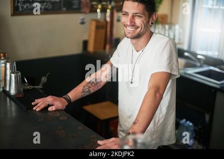 Bereit, Gäste mit einem Lächeln zu begrüßen. Ein glücklicher junger Barkeeper, der hinter dem Tresen in einer Bar steht. Stockfoto