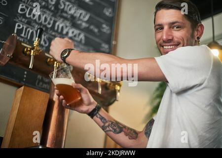 Lust auf eine kalte. Porträt eines jungen Barkeepers, der hinter der Bar Bier vom Zapfhahn gießt. Stockfoto