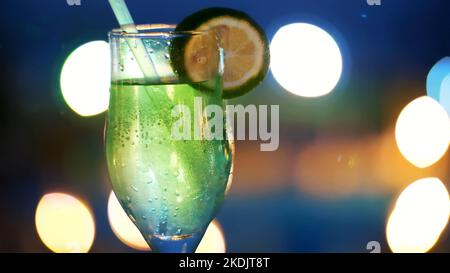 Cocktailparty, trinken Sie mit Eis, in einem Glas mit Stroh und dekoriert mit einer Scheibe Zitrone, in den Strahlen von Soffits, Lampen, flackernde Lichter. Original-alkoholische und alkoholfreie Cocktails. Hochwertige Fotos Stockfoto