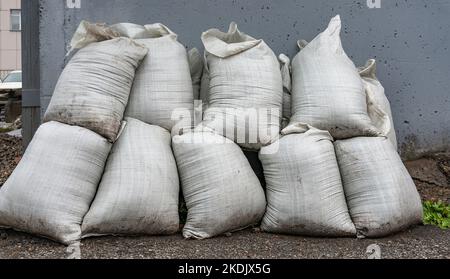 Blick auf einen Haufen weißer Säcke, die auf nassem Asphalt auf der Straße gegen die Wand eines städtischen Gebäudes öffentlicher Versorgungsunternehmen stehen Stockfoto