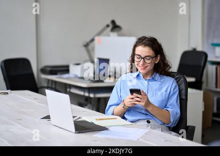Junge Geschäftsfrau, die Mobiltelefon benutzt und im Büro am Schreibtisch sitzt. Stockfoto