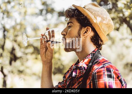 Hübscher bärtiger Kerl, der beim Weinerntepicknick unter Bäumen und Weinreben Rotwein trinkt - Essen und Trinken - Lifestyle-Konzept der Menschen Stockfoto