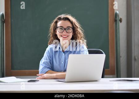 Junge glückliche weibliche Online-Lehrerin, die mit einem Laptop am Schreibtisch sitzt. Hochformat Stockfoto