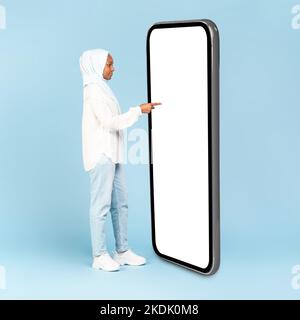 Schwarze muslimische Frau mit großem Handy mit leerem Bildschirm und berühren riesige Display-Panel mit Finger, Mockup Stockfoto