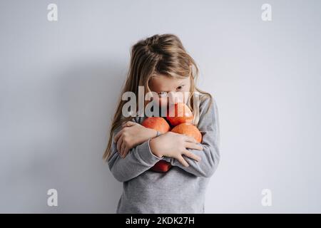 Ein wehmütiger Junge umarmte einen Haufen Äpfel in seinen Armen. Über einer weißen Wand. Er hat lange blonde Haare. Stockfoto