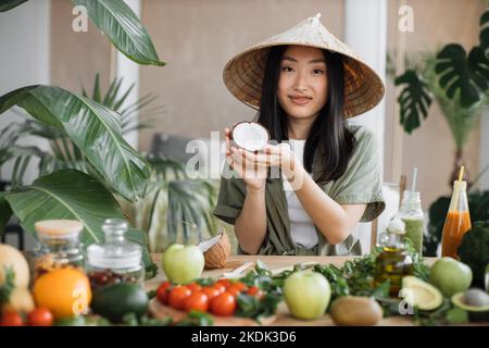 Schöne asiatische Frau in traditionellen konischen Hut hält Kokosnuss sitzen am Tisch mit Obst und Gemüse Zutaten zu gesunden veganen Salat, drinnen am Tisch in der stilvollen tropischen Küche. Stockfoto