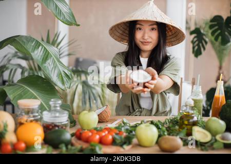 Schöne asiatische Frau in traditionellen konischen Hut hält Kokosnuss sitzen am Tisch mit Obst und Gemüse Zutaten zu gesunden veganen Salat, drinnen am Tisch in der stilvollen tropischen Küche. Stockfoto