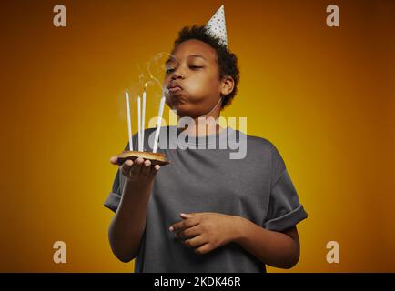 Süßer afroamerikanischer Youngster mit Geburtstagskappe und grauem T-Shirt, der brennende Kerzen bläst und sich im Studio seinen Geburtstag wünscht Stockfoto