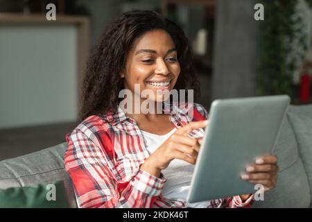 Frohes glückliches schwarzes junges Weib, das gute Nachricht liest, auf Tablet tippt, Video im Wohnzimmer innen anschaut Stockfoto