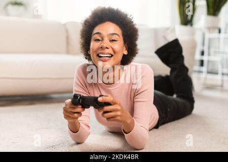 Fröhliche tausendjährige afroamerikanische Frau mit Joystick auf dem Boden liegend, Online-Spiele im Wohnzimmer zu spielen Stockfoto
