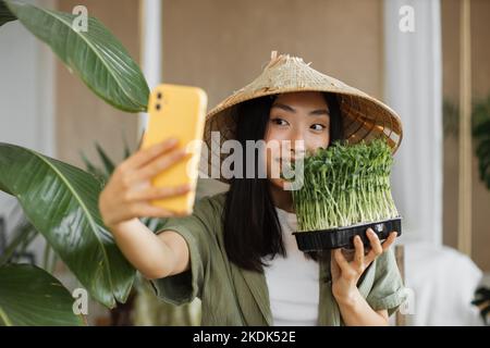 Schöne asiatische Frau Bloggerin in traditionellen Hut Herstellung von Gemüse Obst und mikrogrünen Sprossen Salat und machen Video für Social Media genießen gesundes Abendessen in exotischen Licht Studio im Resort Stockfoto