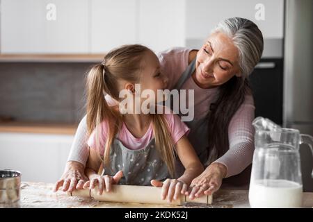 Fröhliche hübsche kaukasische kleine Enkelin und ältere Großmutter erzählen Teig mit Nudelholz zum Backen Stockfoto