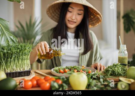 Junge attraktive asiatische Frau in traditionellen konischen Hut sitzt am Tisch in der stilvollen hellen tropischen Küche und gießt Olivenöl zu gesunden Gemüsesalat. Stockfoto