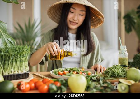 Junge attraktive asiatische Frau in traditionellen konischen Hut sitzt am Tisch in der stilvollen hellen tropischen Küche und gießt Olivenöl zu gesunden Gemüsesalat. Stockfoto