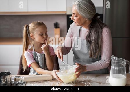 Glückliche kaukasische kleine Enkelin und reife Großmutter mit Mehl beschmiert vorbereiten, schmecken Teig zum Backen Stockfoto