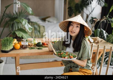 Schöne glückliche junge asiatische Frau in traditionellen konischen Hut hält und essen gesunden Salat sitzen am Stuhl neben dem Tisch mit Bio-Gemüse und Obst Zutaten drinnen in tropischen Resort. Stockfoto