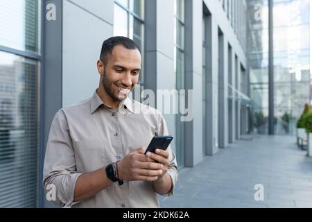 hispanischer Mann vor dem modernen Bürogebäude mit Smartphone, Geschäftsmann im Hemd, der Nachrichten tippt und Online-Seiten durchsucht. Stockfoto