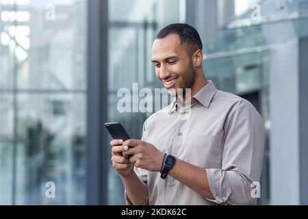 hispanischer Mann vor dem modernen Bürogebäude mit Smartphone, Geschäftsmann im Hemd, der Nachrichten tippt und Online-Seiten durchsucht. Stockfoto