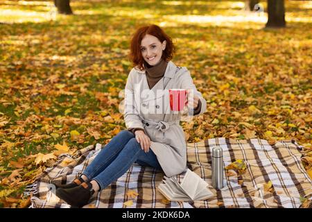 Lächelndes, tausendjähriges kaukasisches Weibchen mit roten Haaren im Regenmantel sitzt auf karierten Thermoskannen Stockfoto